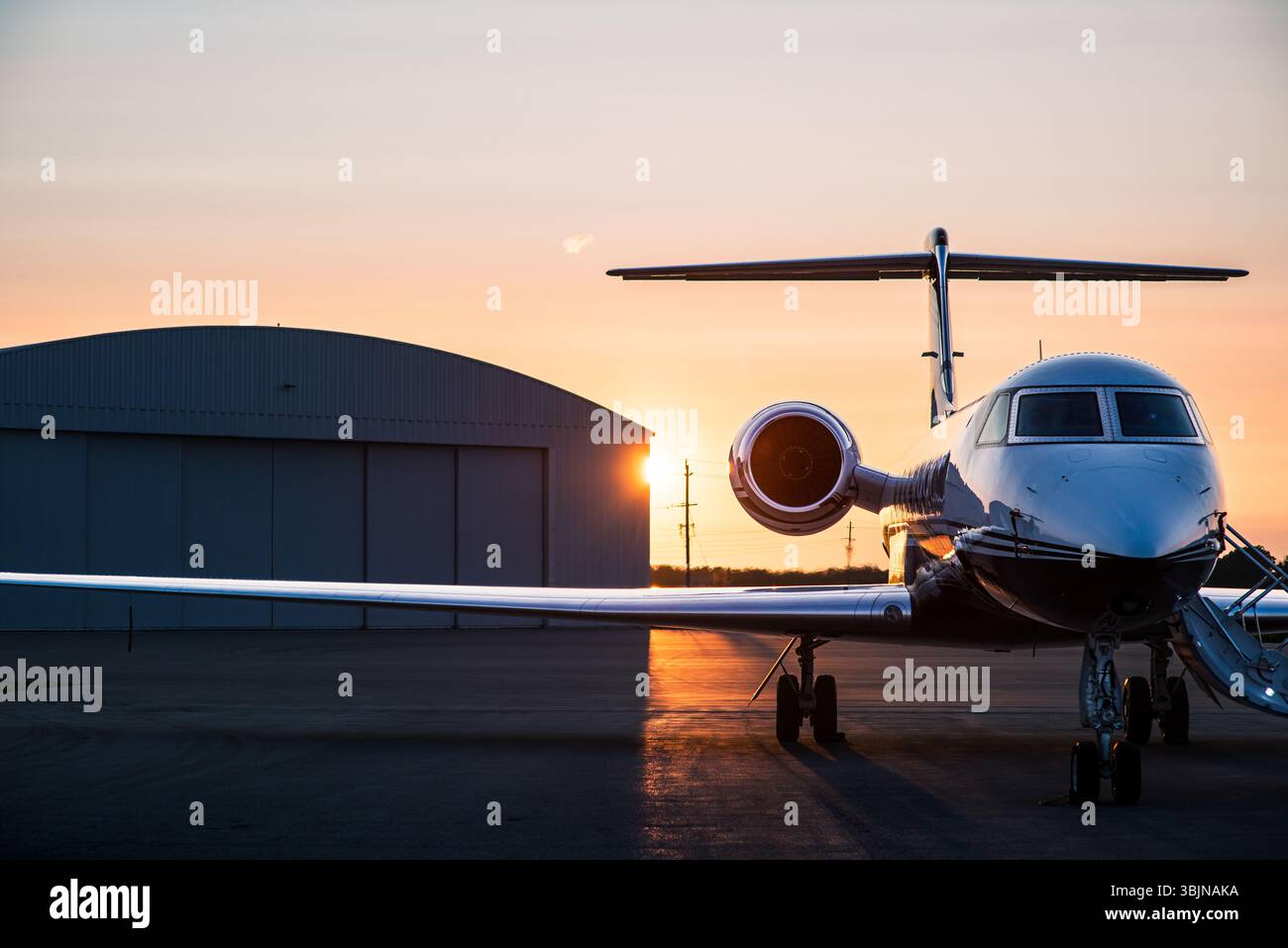 Jet privé silhouetté contre un coucher de soleil doré, garé sur le tarmac à côté d'un hangar. Départs en soirée et voyages de luxe. Banque D'Images