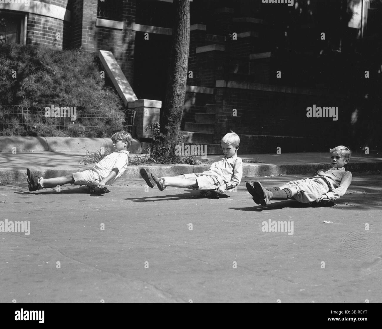 Trois enfants assis sur des planches à roulettes, années 1920, trois garçons jouant sur des planches à roulettes dans une rue de la ville, photo historique Banque D'Images
