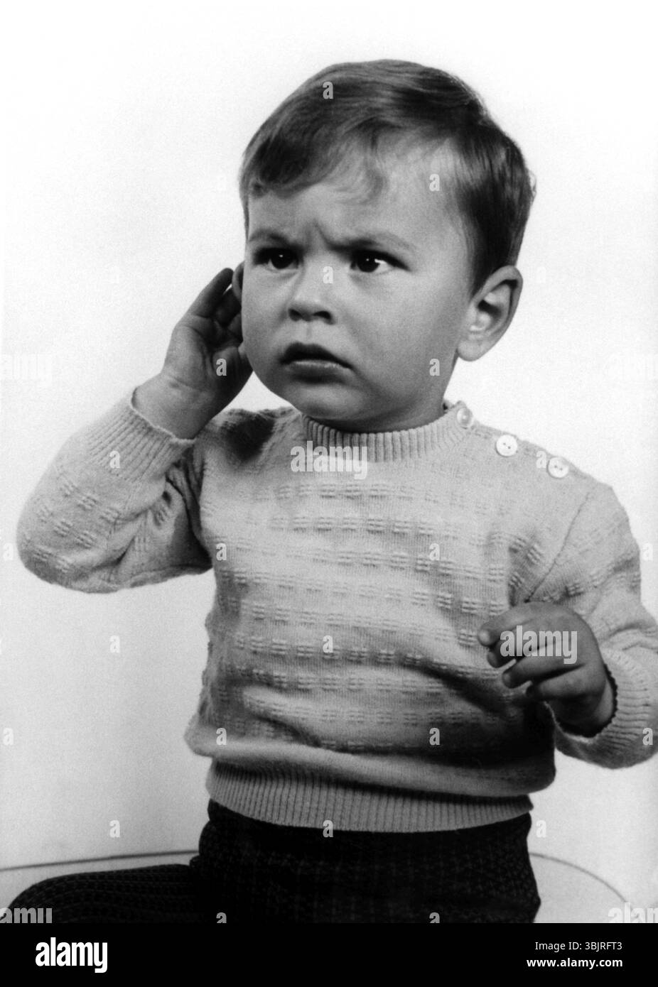 Enfant agacé, des années 1950, Un petit garçon dans un pull avec une expression questionnant tient sa main à son oreille, photo historique Banque D'Images