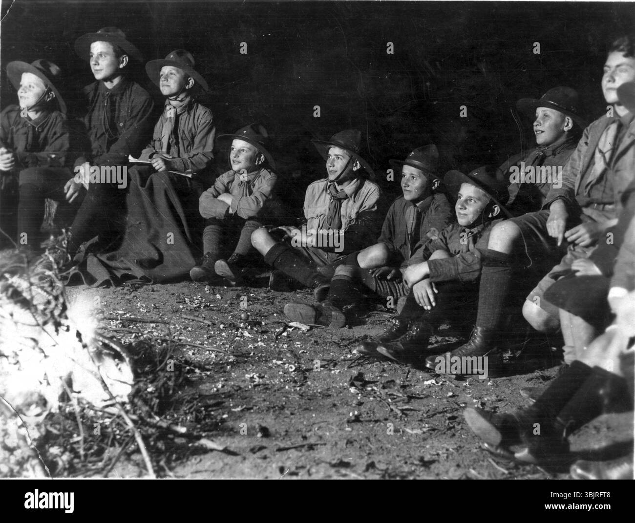 Feu de camp scout, années 1930, Groupe de garçons en costumes scout assis attentivement autour d'un feu de camp dans l'obscurité, photo historique Banque D'Images