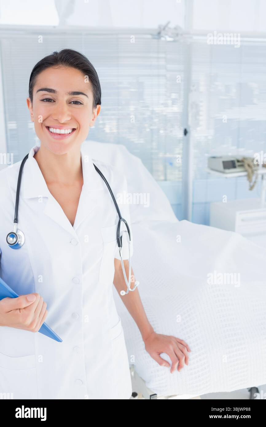 Médecin debout à côté du lit du patient dans une salle clinique lumineuse, avec presse-papiers et stéthoscope Banque D'Images