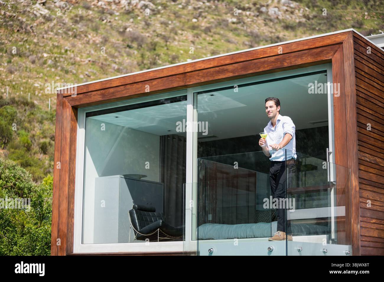 homme debout sur un balcon à balustrade de verre tenant un verre surplombant une colline herbeuse, à côté d'une maison en bois Banque D'Images