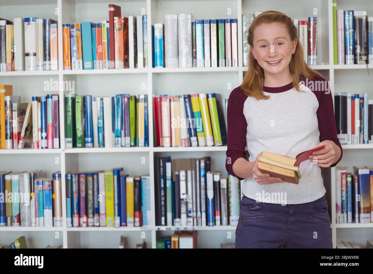Enfant femelle lisant un livre ouvert tout en se tenant debout près de l'étagère de la bibliothèque avec des livres à couverture rigide, espace de copie Banque D'Images