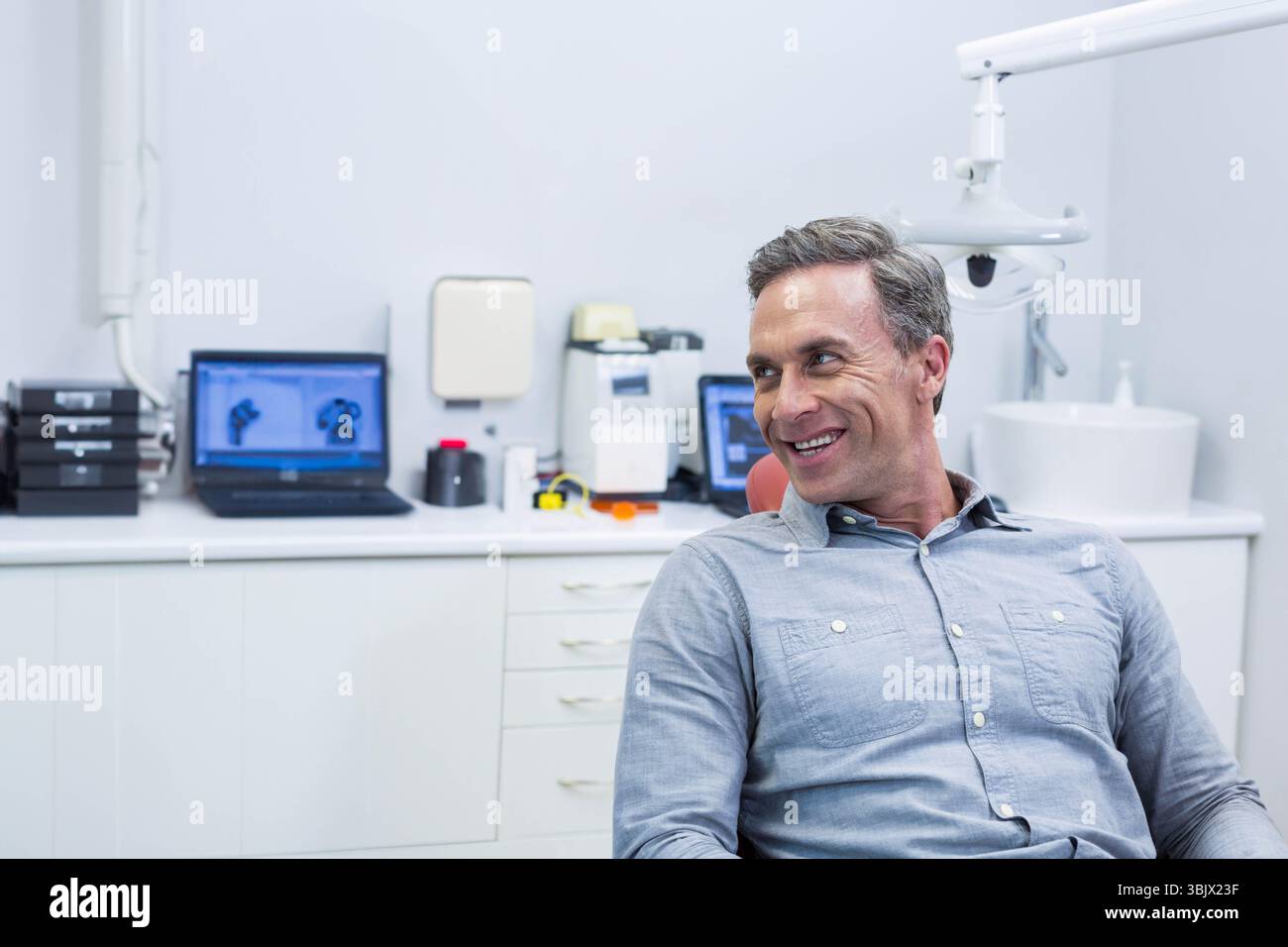 Homme d'âge moyen assis dans un fauteuil opératoire à la clinique dentaire avec des ordinateurs portables, lampe plafonnière, espace de copie Banque D'Images