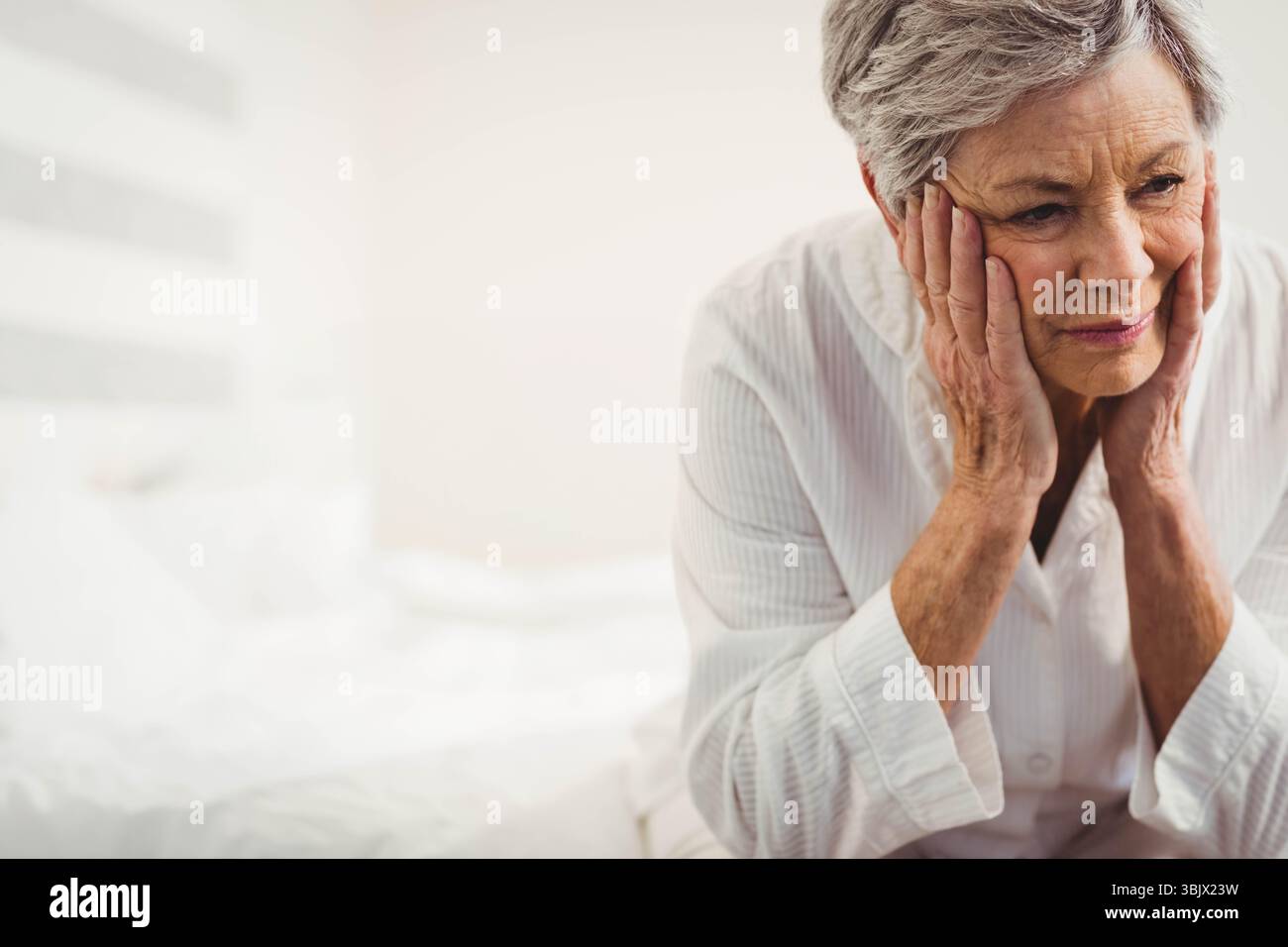 Femme senior assise sur le lit de la chambre avec des oreillers blancs portant pyjama pressant les joues, espace de copie Banque D'Images