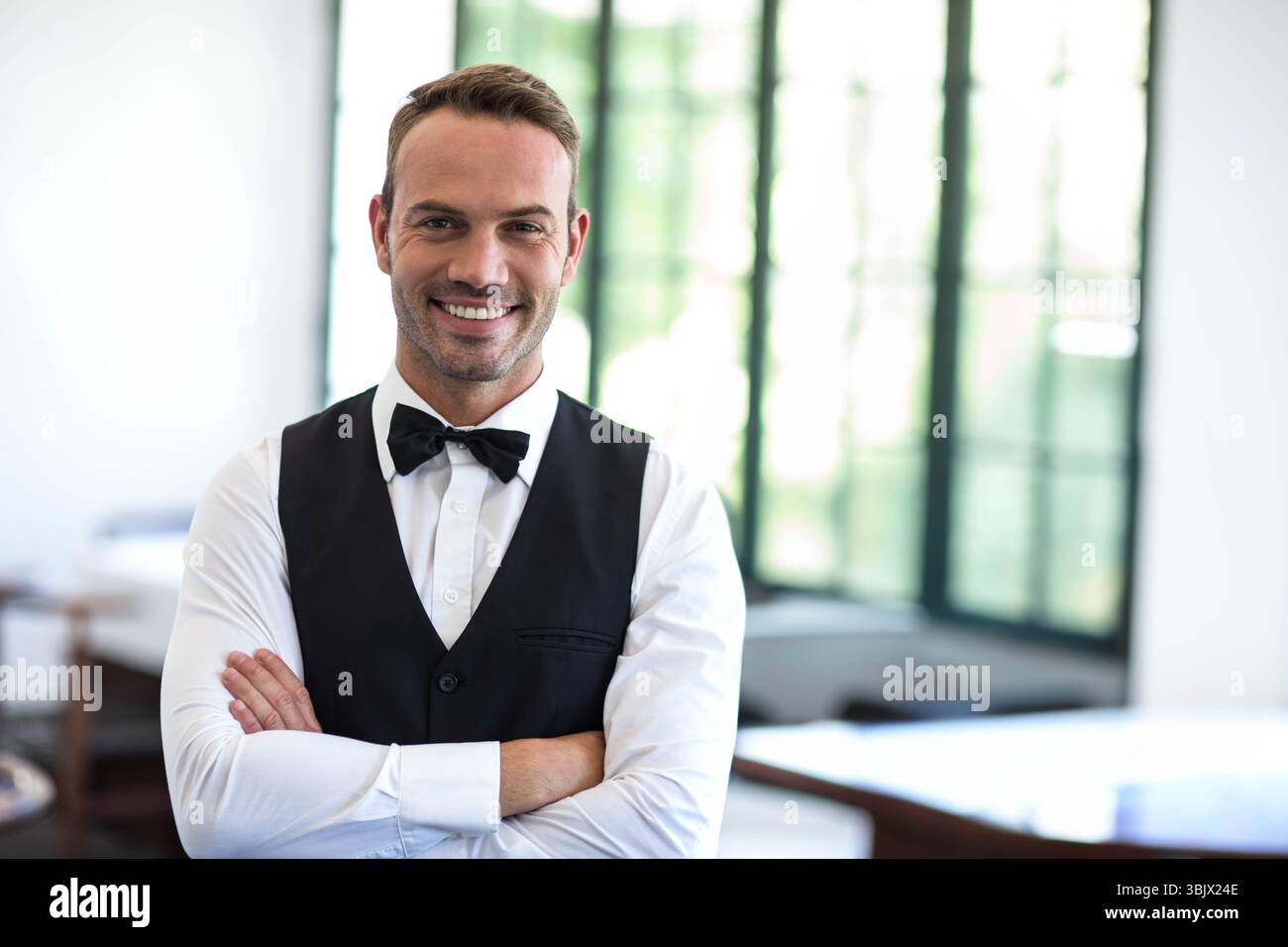 Garçon adulte moyen portant des bras croisés uniformes souriant dans la salle à manger avec tables et fenêtre Banque D'Images