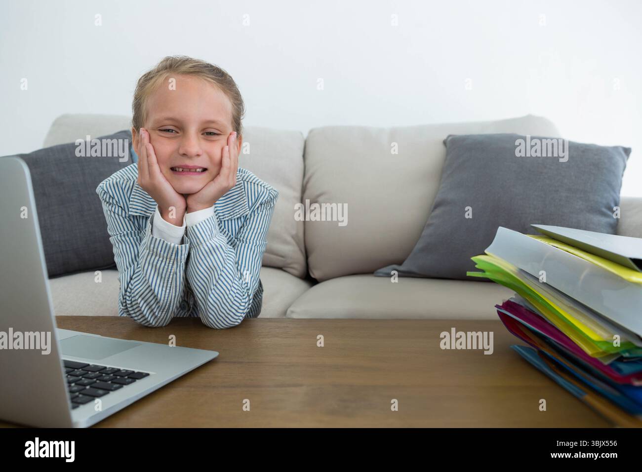Fille menton reposant sur les mains souriant à la table en bois avec ordinateur portable argenté et dossiers colorés Banque D'Images