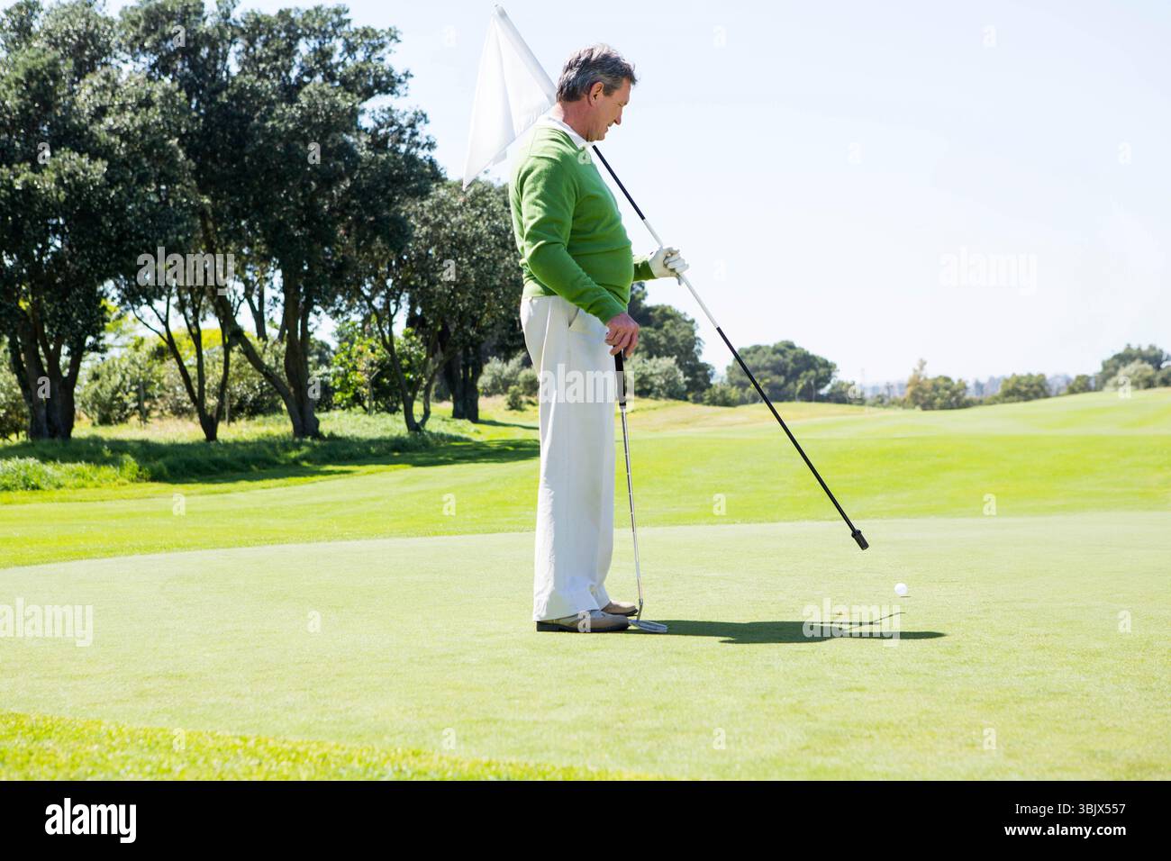 Golfeur senior tenant un drapeau et un putter sur un vert bien entretenu, avec une balle près du trou Banque D'Images