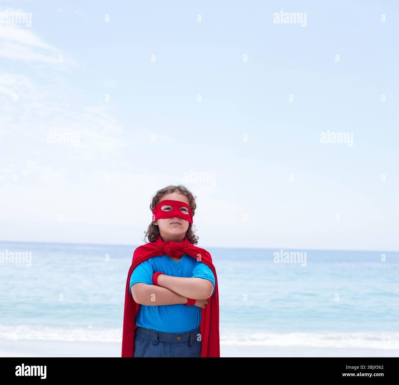 L'enfant est debout sur la plage de sable portant un masque de super-héros rouge, une cape et un short, face à l'horizon de l'océan Banque D'Images