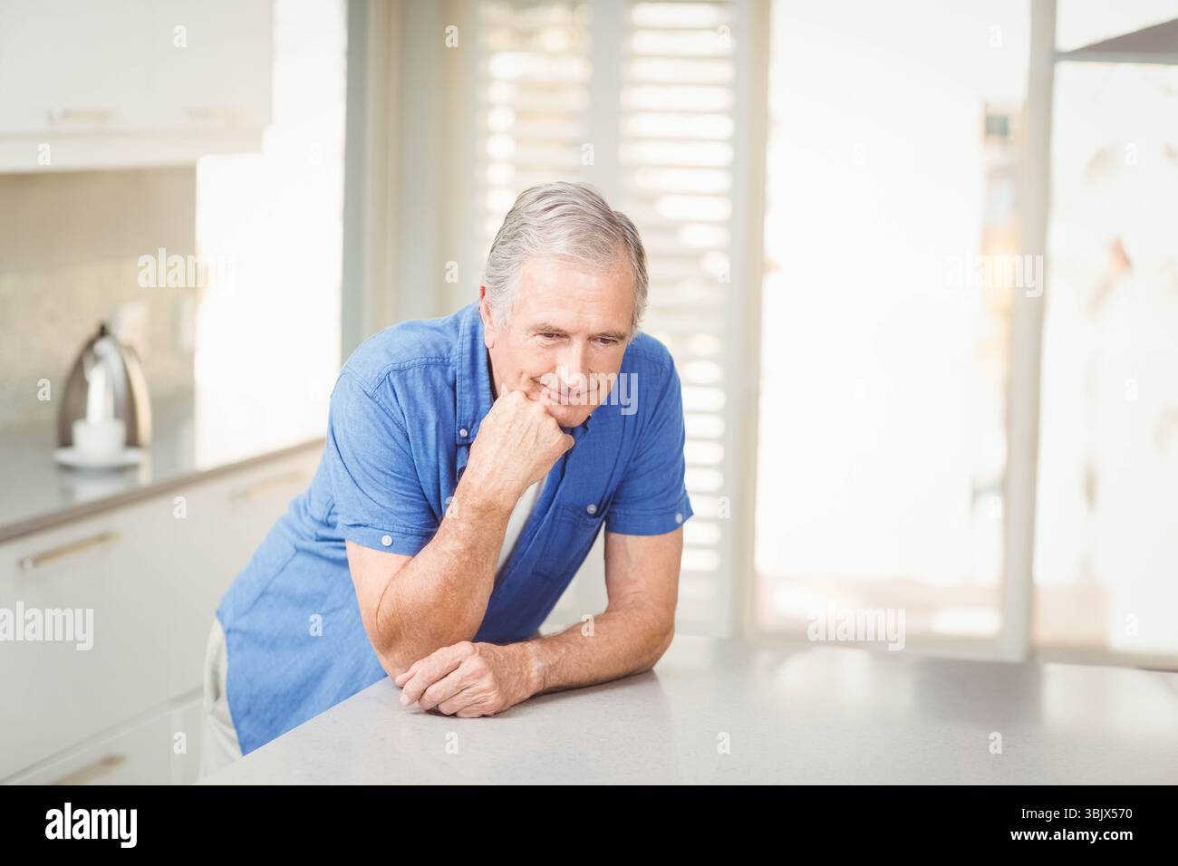 Homme senior appuyé sur le comptoir de quartz dans la cuisine moderne avec bouilloire et volets Banque D'Images