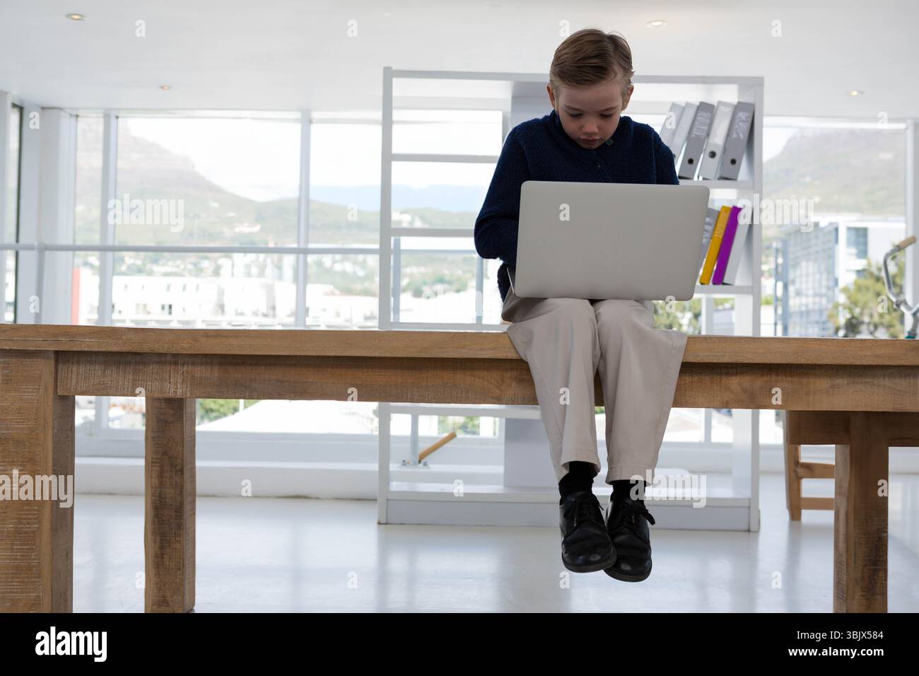 Garçon enfant assis sur le bord de la table dans l'étude lumineuse à l'aide d'un ordinateur portable argenté près des classeurs à anneaux Banque D'Images