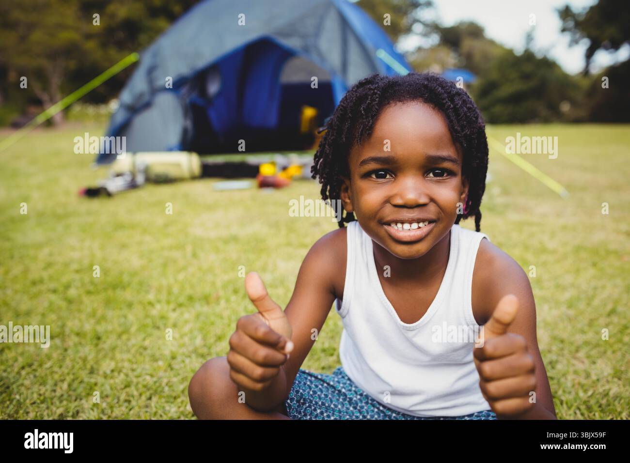 Garçon afro-américain assis sur un champ herbeux devant la tente bleue donnant les pouces avec un sac de couchage Banque D'Images