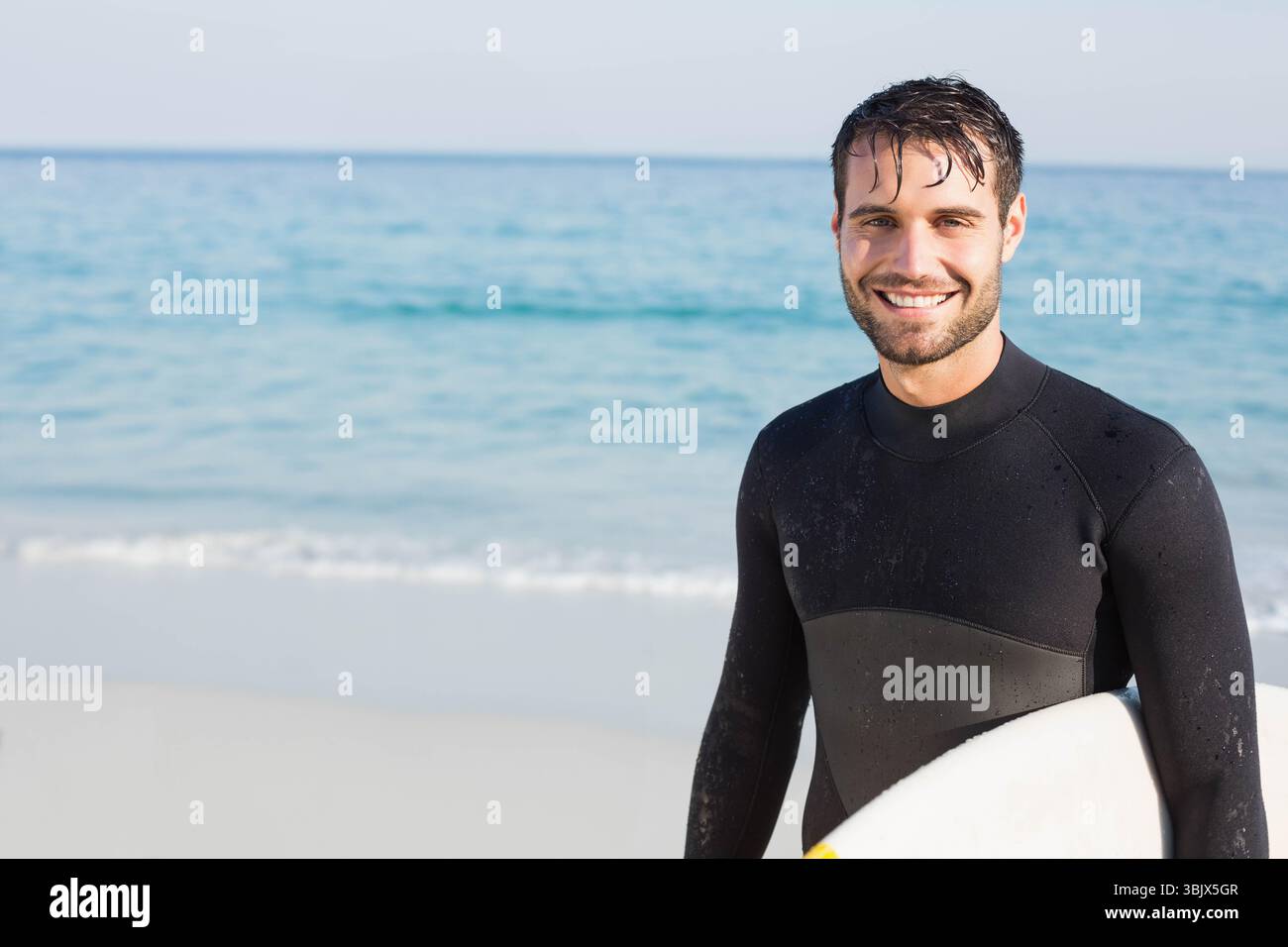 Homme tenant une planche de surf et souriant au bord de la plage portant une combinaison noire face à l'océan, espace copie Banque D'Images