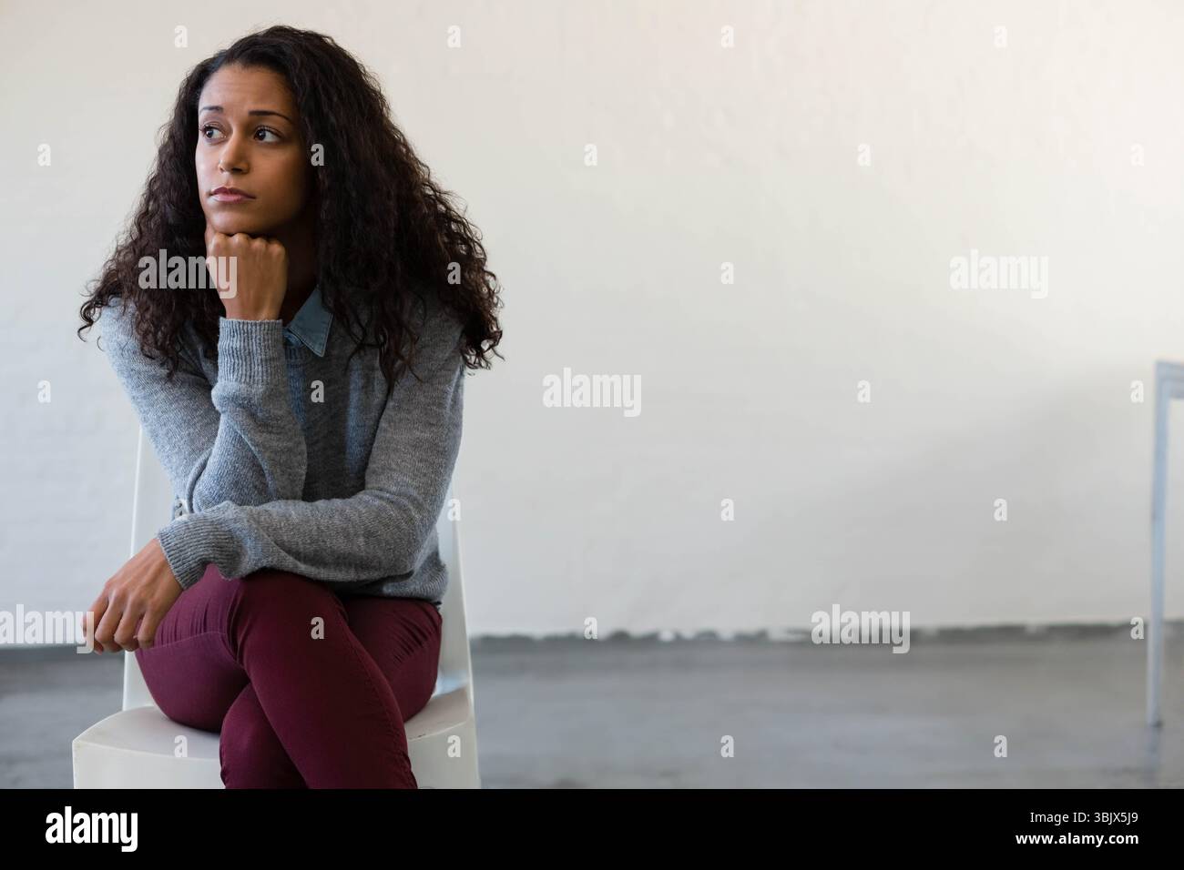 Femme assise pensivement sur une chaise blanche en studio portant un pull gris avec pied de table, espace copie Banque D'Images