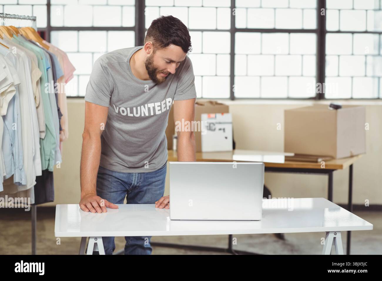 Homme volontaire penché sur la table pliante tapant sur l'ordinateur portable dans l'entrepôt à côté du rack de chemises Banque D'Images