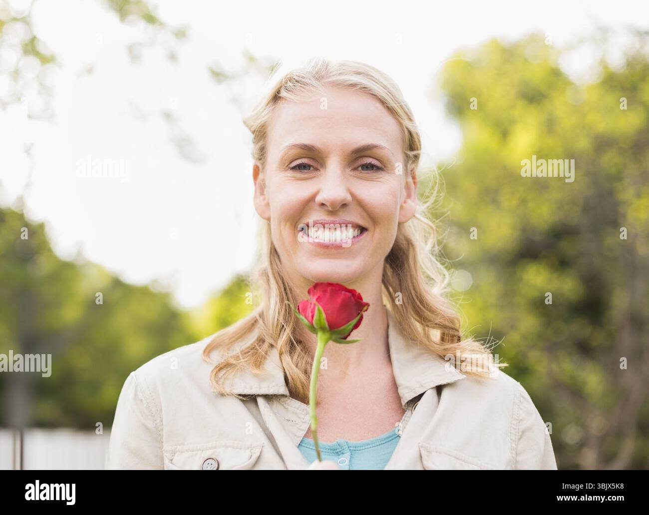 Femme adulte moyenne tenant une rose rouge simple et souriante à l'extérieur dans le jardin avec feuillage vert Banque D'Images