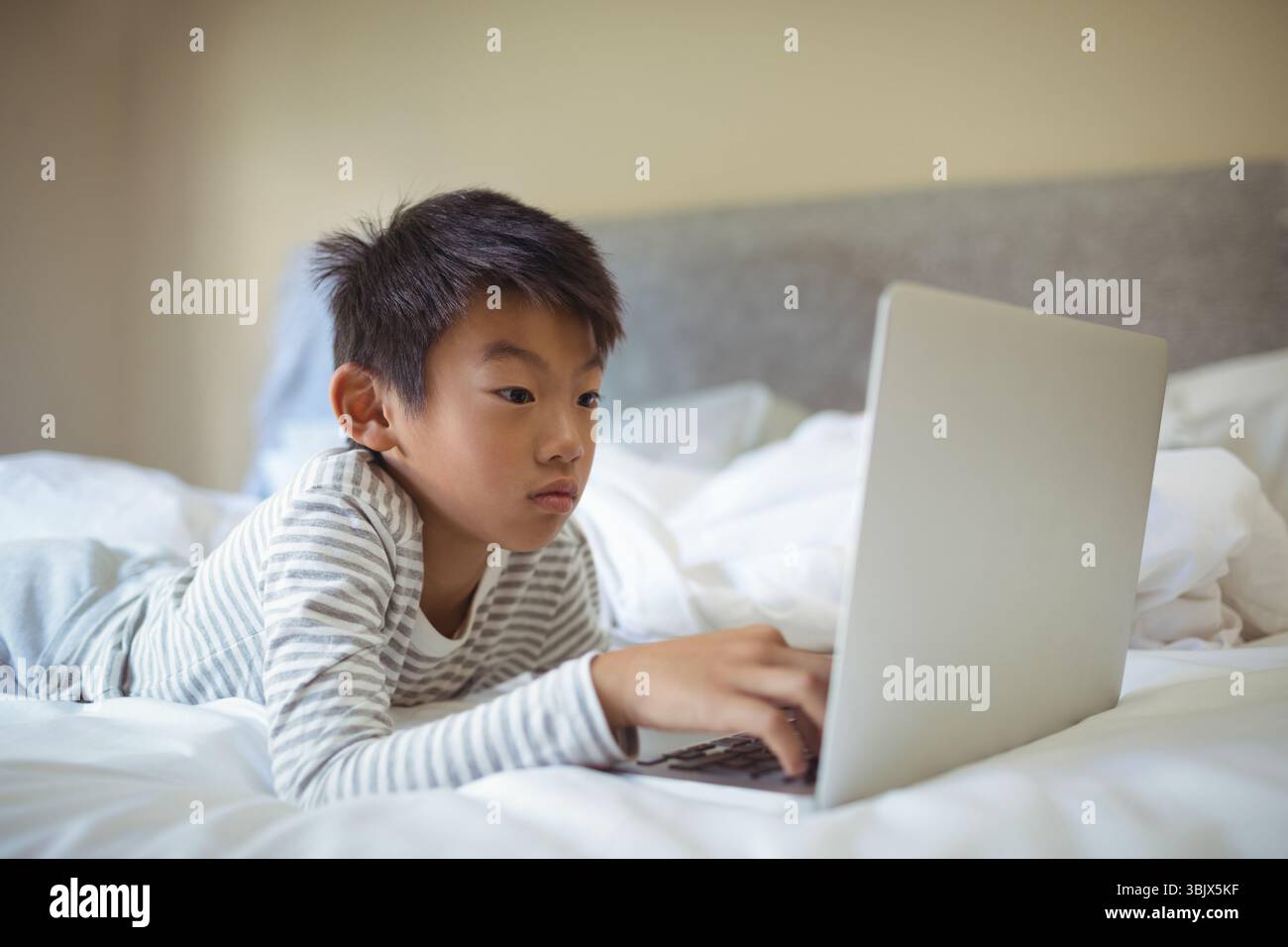 Garçon d'enfant asiatique allongé sur le lit en utilisant un ordinateur portable argenté sur la literie blanche grondée dans la chambre Banque D'Images