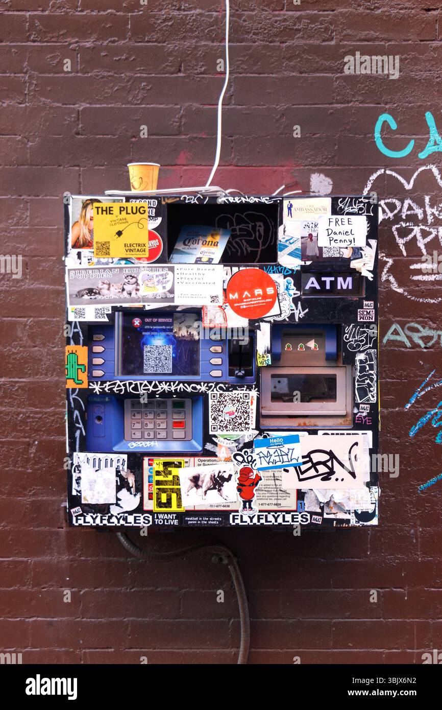 Un distributeur vintage recouvert de graffitis et d'autocollants installés sur un mur de briques avec du Street art urbain et des motifs colorés qui l'entourent Banque D'Images