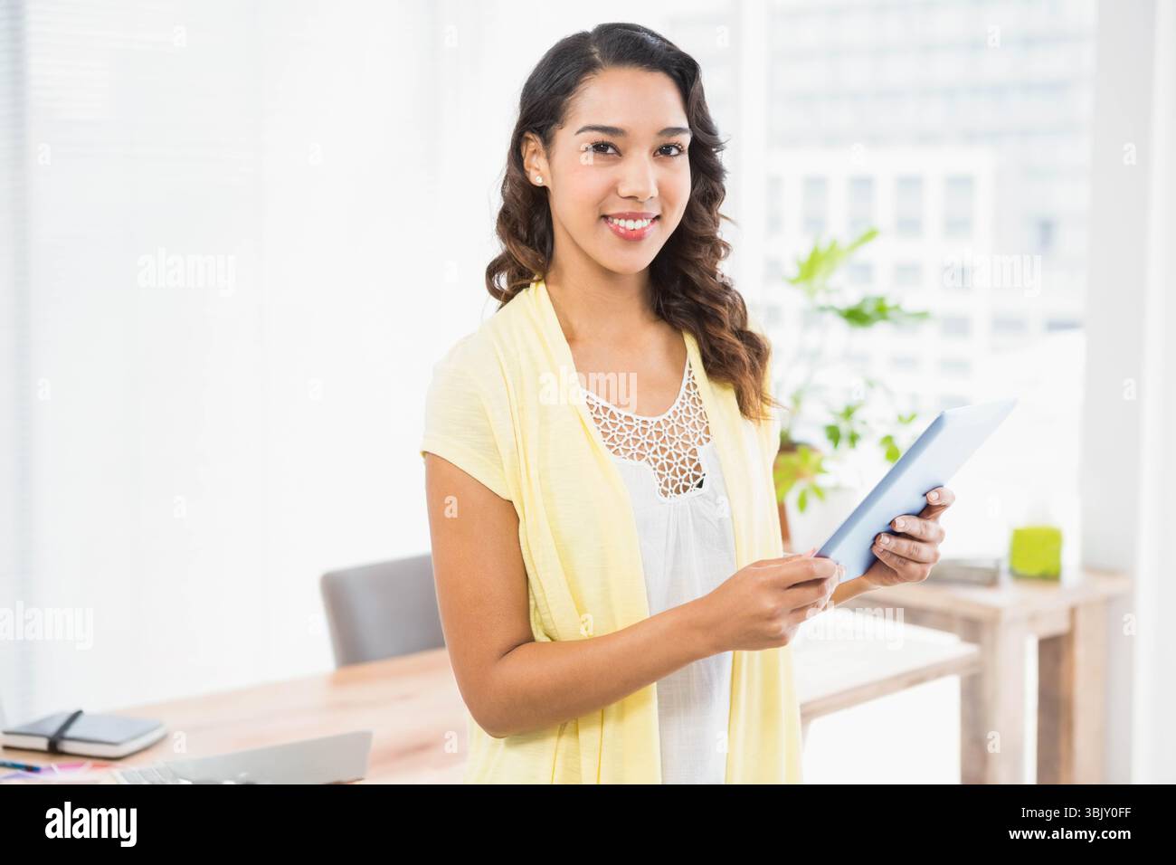 Bureau en bois affichant un ordinateur portable fermé, une tablette et un ordinateur portable à côté d'une plante feuillue près d'une grande fenêtre Banque D'Images