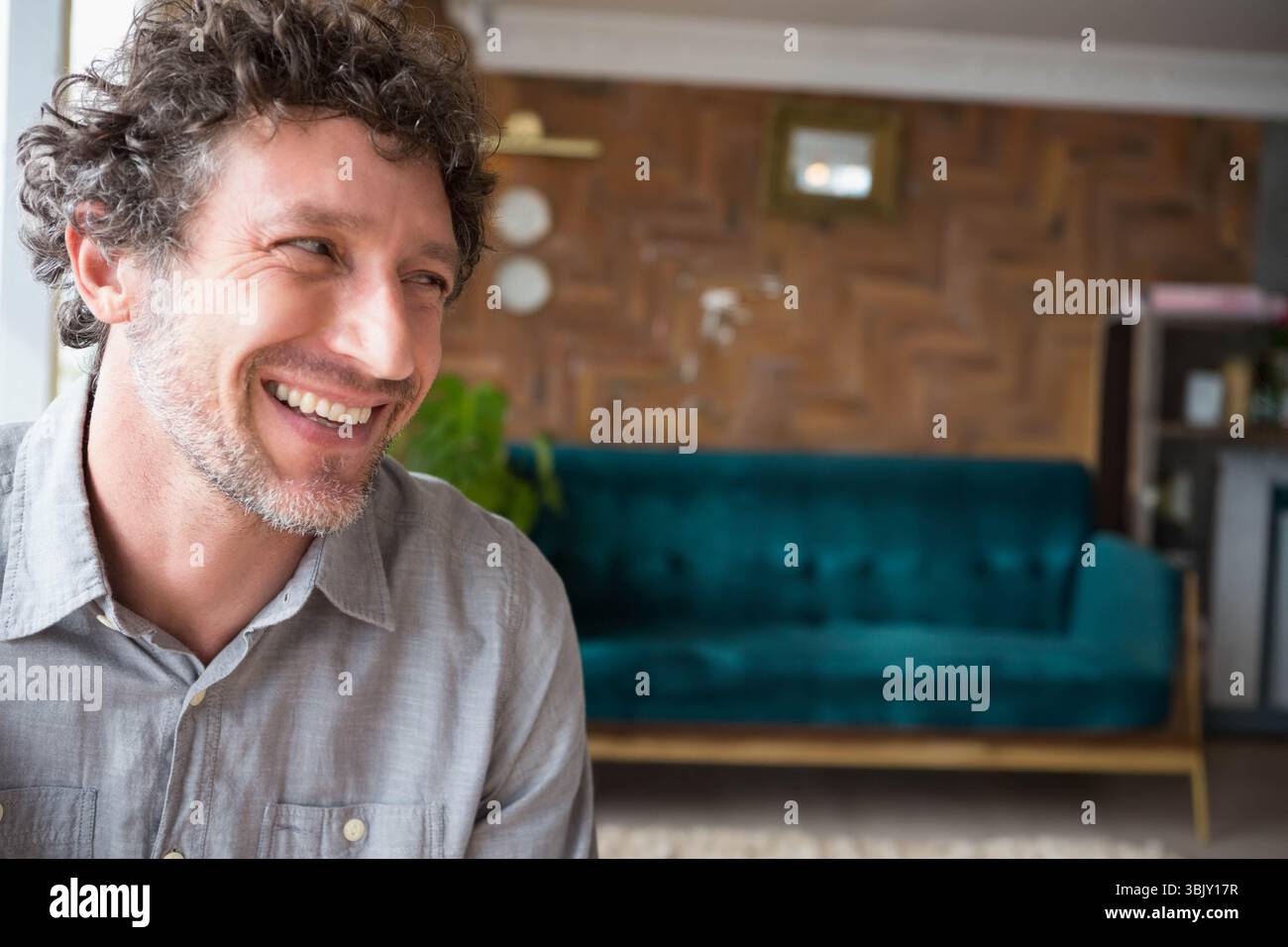Homme d'âge moyen souriant et assis dans le salon avec canapé sarcelle, plante en pot et étagère Banque D'Images