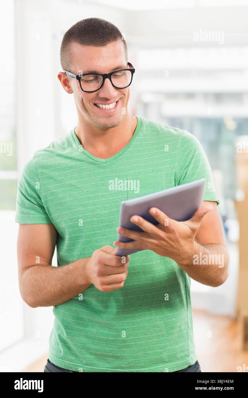 Homme en T-shirt rayé vert souriant et tapotant tablette dans l'espace de travail lumineux près des fenêtres Banque D'Images