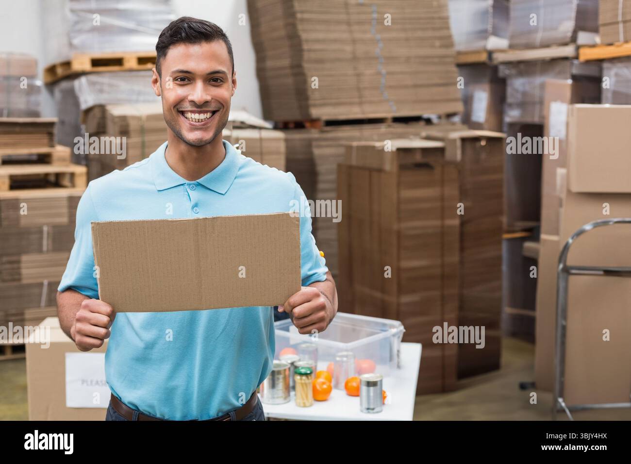 Ouvrier d'entrepôt masculin tenant le signe de carton vierge dans l'entrepôt au milieu des boîtes aplaties et du conteneur Banque D'Images