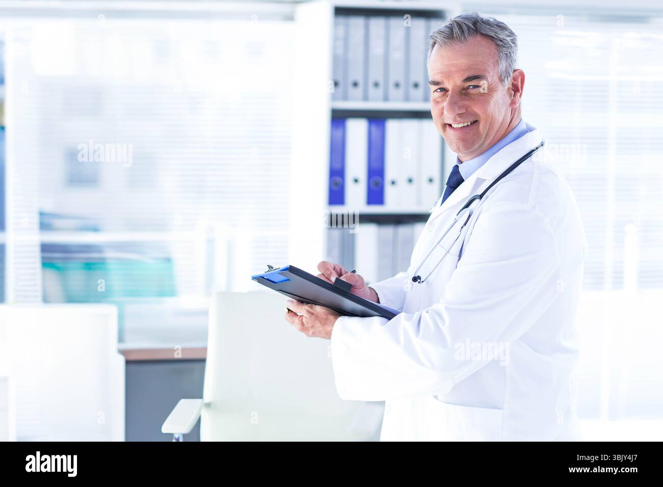 Médecin masculin écrivant presse-papiers avec stylo dans le stéthoscope de blouse de laboratoire à la clinique par étagère, espace de copie Banque D'Images