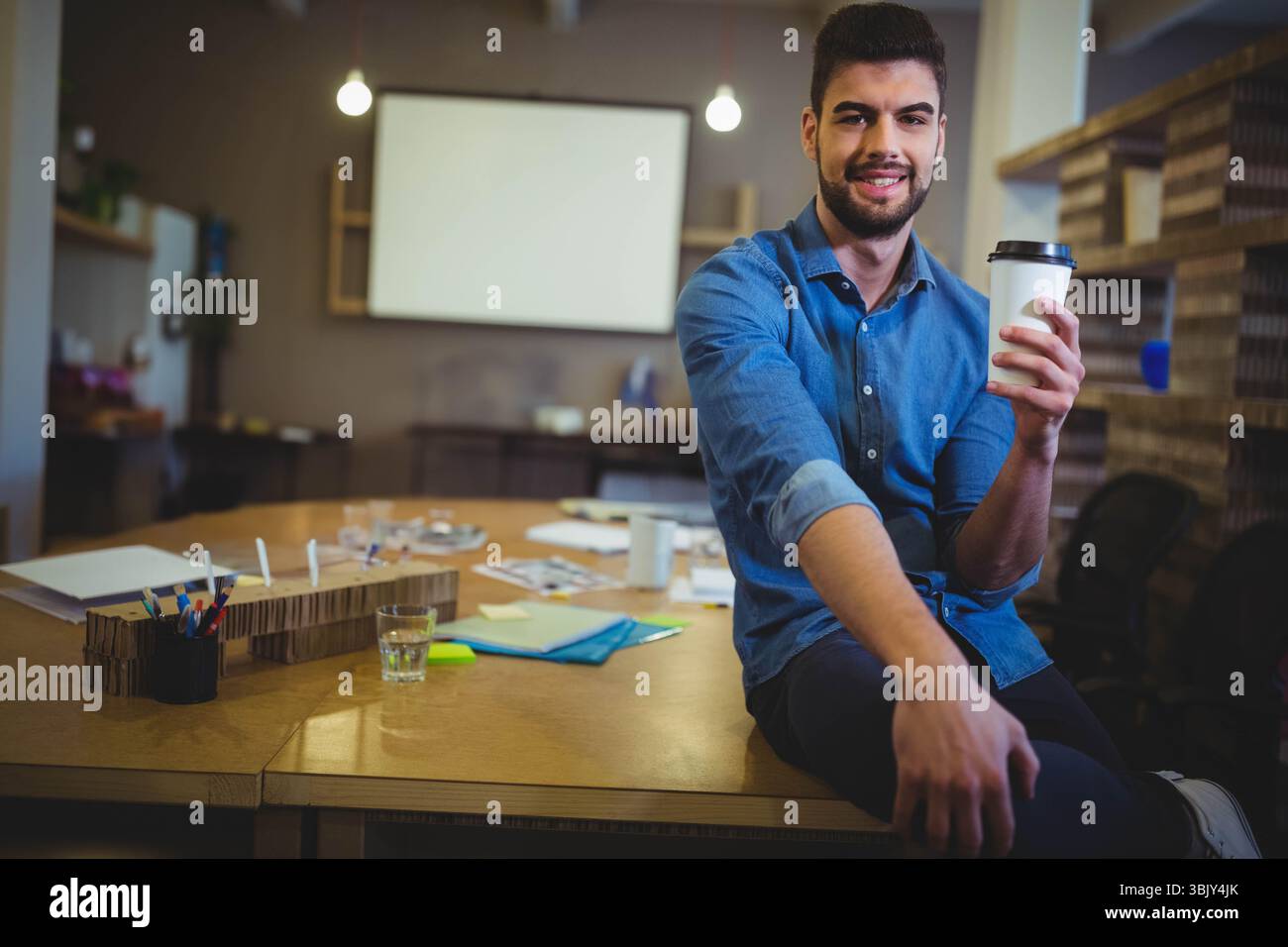 Homme assis sur le bord du bureau dans le bureau sirotant un café avec un écran de projecteur vierge derrière, espace de copie Banque D'Images