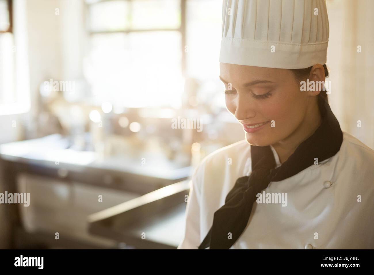 Femme chef debout dans la cuisine portant un manteau blanc et chapeau plissé par comptoir en acier, espace de copie Banque D'Images