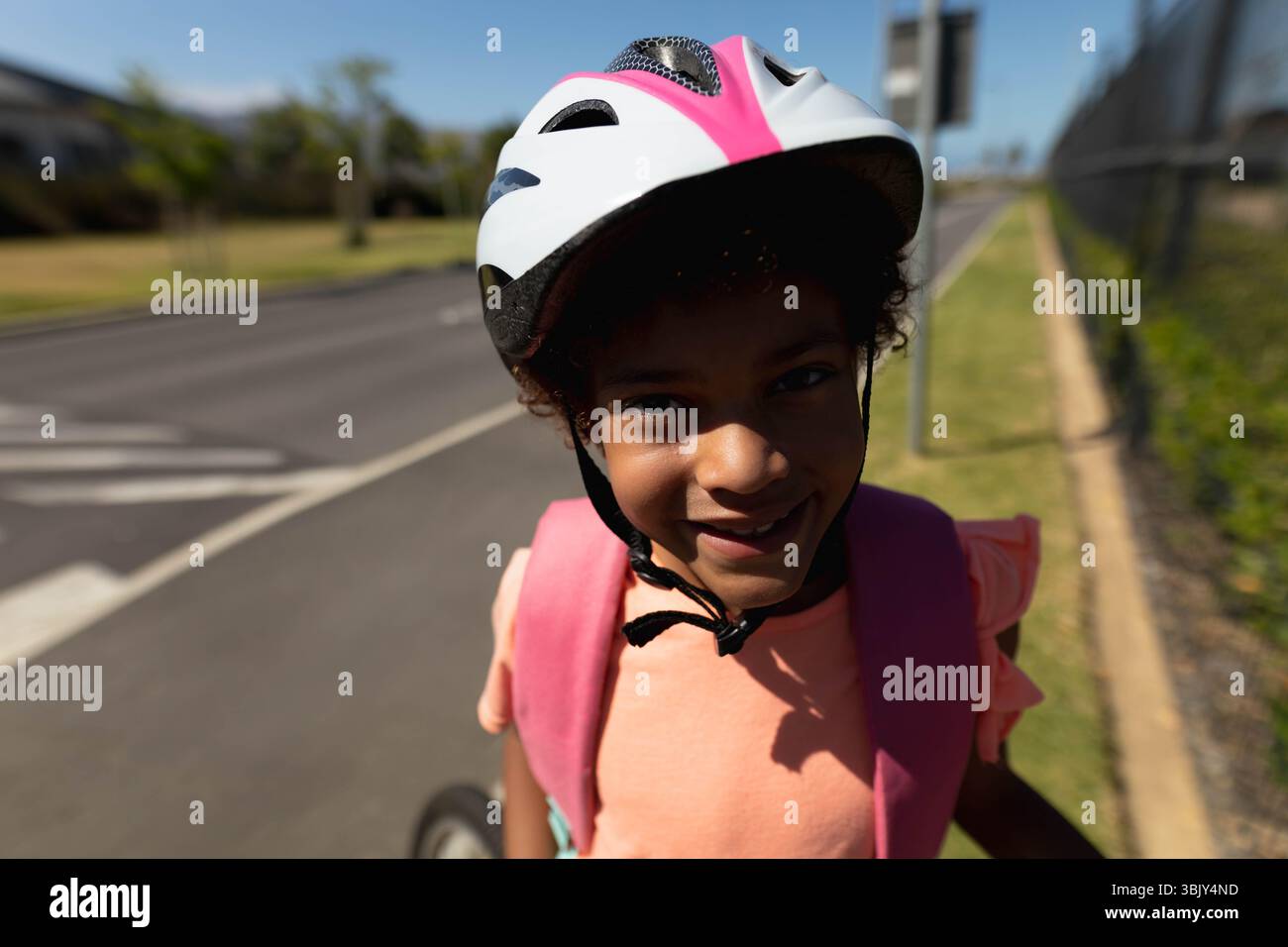 Fille afro-américaine debout sur le trottoir de banlieue portant un casque de vélo blanc-rose et un sac à dos Banque D'Images
