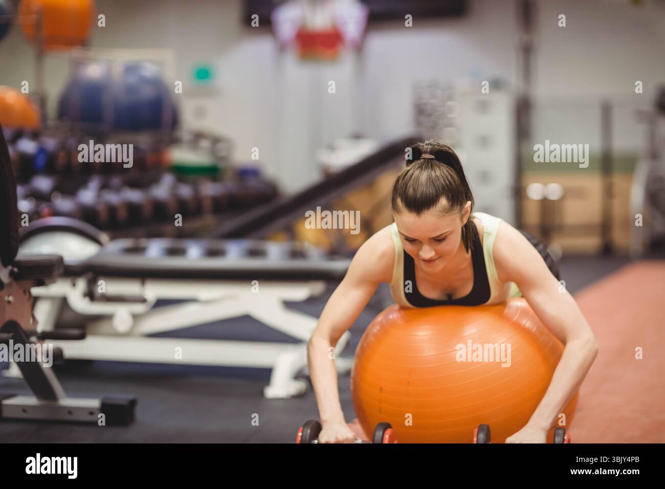 Routine d'entraînement de stabilité féminine à la salle de gym en s'appuyant sur un ballon d'équilibre orange et en saisissant des haltères Banque D'Images