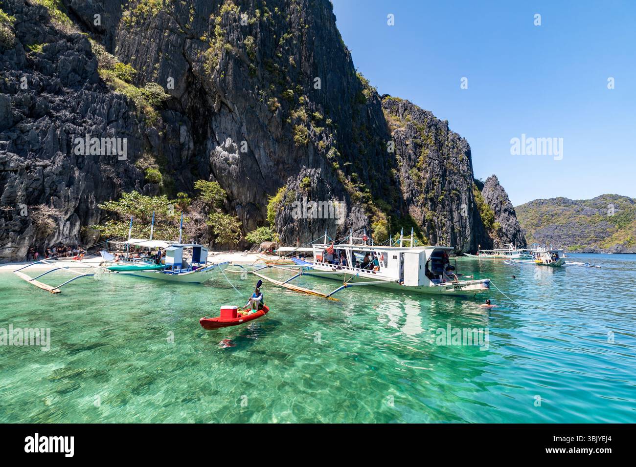 Une personne fait du kayak dans les eaux claires d'El Nido, aux Philippines, près de plusieurs bateaux et d'une plage avec des gens, profitant probablement d'une journée d'île en île et d'activités nautiques. Banque D'Images