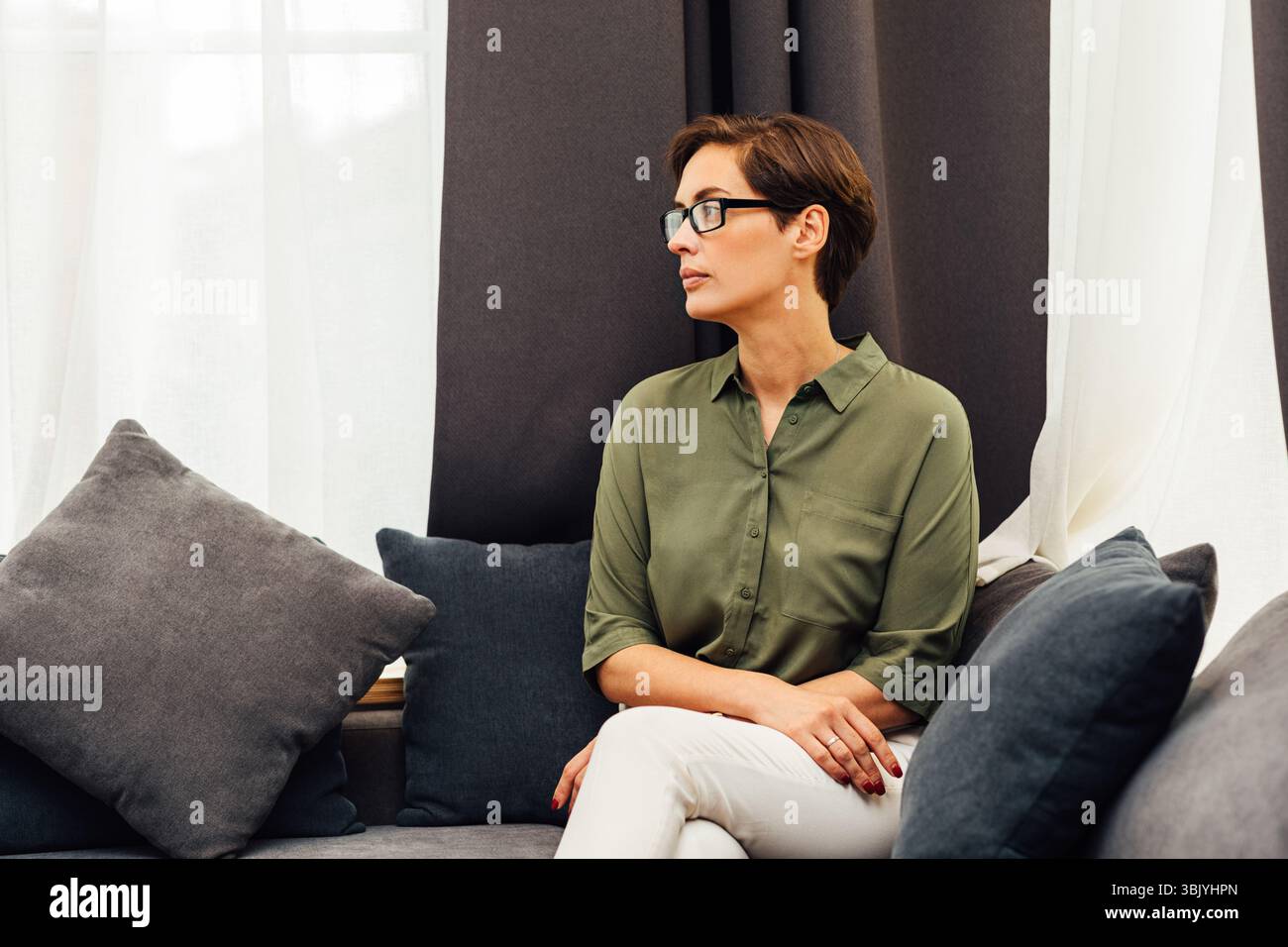Vue latérale d'une femme élégante assise sur un canapé dans la chambre d'hôtel et vue sur l'extérieur Banque D'Images Vue latérale d'une femme élégante assise sur un canapé dans la chambre d'hôtel et vue sur l'extérieur Banque D'Images