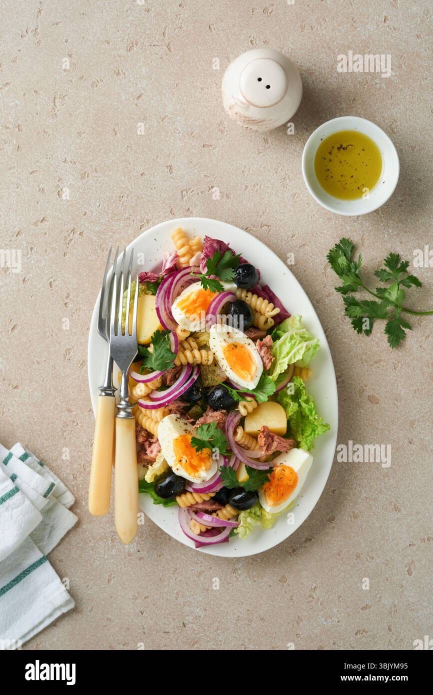 Salade de thon avec pâtes, œufs, pommes de terre, olives, oignons rouges et sauce dans une assiette blanche sur fond de table en béton gris clair vieux. Banque D'Images Salade de thon avec pâtes, œufs, pommes de terre, olives, oignons rouges et sauce dans une assiette blanche sur fond de table en béton gris clair vieux. Banque D'Images