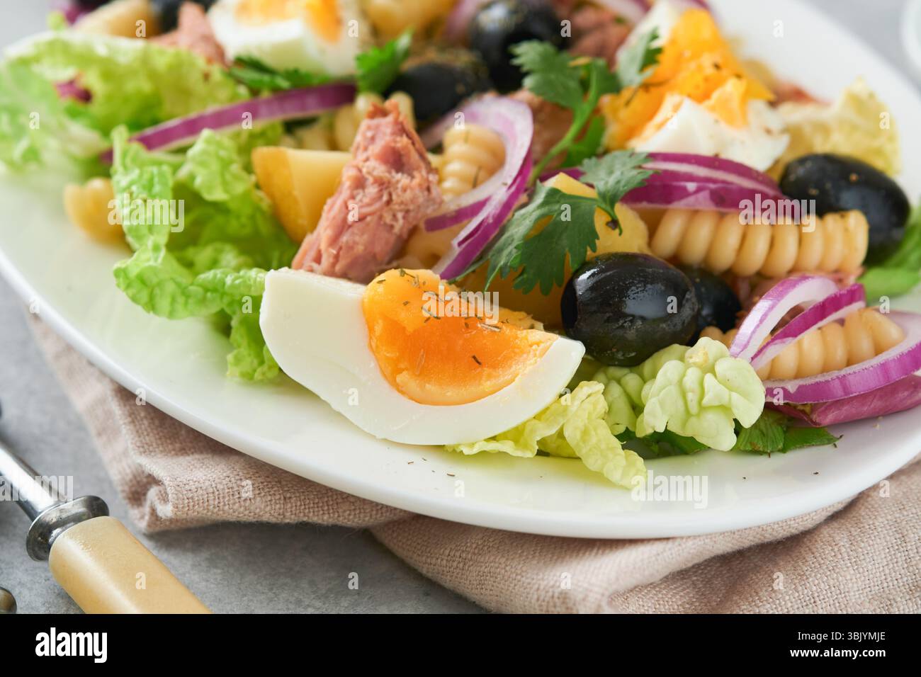 Salade de thon avec pâtes, œufs, pommes de terre, olives, oignons rouges et sauce dans une assiette blanche sur fond de table en béton gris clair vieux. Banque D'Images Salade de thon avec pâtes, œufs, pommes de terre, olives, oignons rouges et sauce dans une assiette blanche sur fond de table en béton gris clair vieux. Banque D'Images