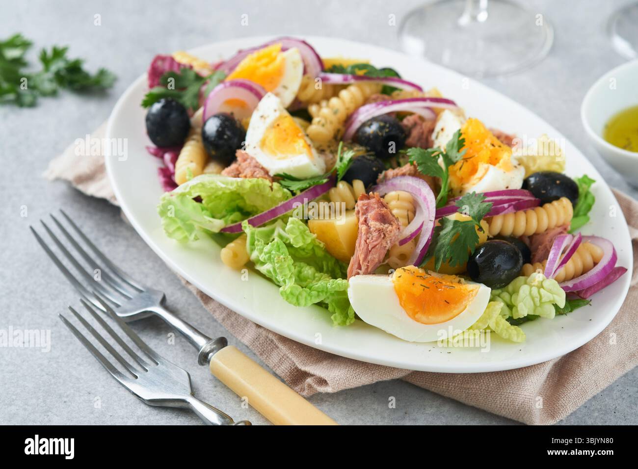 Salade de thon avec pâtes, œufs, pommes de terre, olives, oignons rouges et sauce dans une assiette blanche sur fond de table en béton gris clair vieux. Banque D'Images Salade de thon avec pâtes, œufs, pommes de terre, olives, oignons rouges et sauce dans une assiette blanche sur fond de table en béton gris clair vieux. Banque D'Images