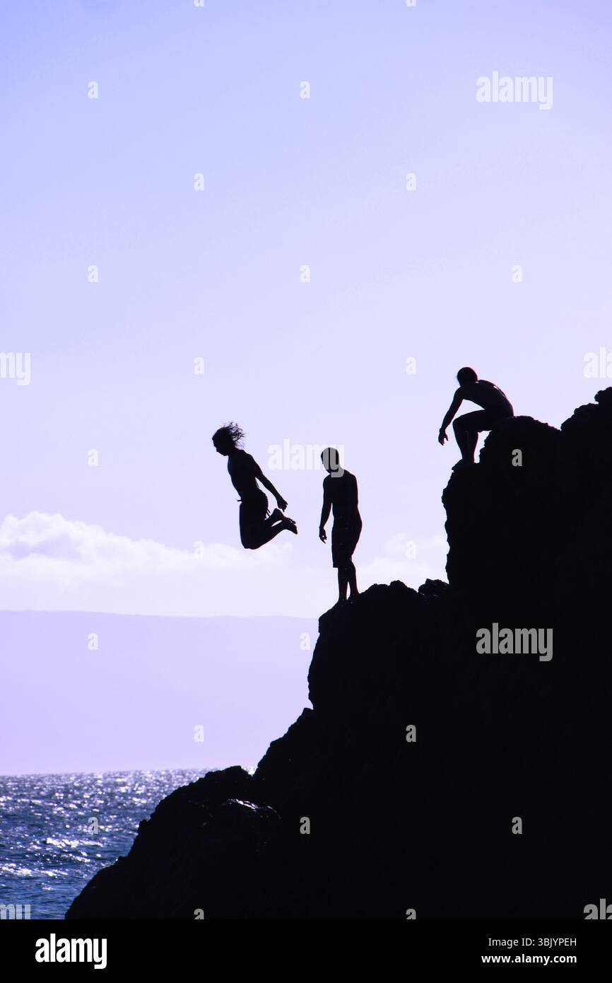 Silhouetted figures falaise plongeant dans l'océan depuis Black Rock à Maui, Hawaï, avec une personne en plein air et d'autres attendant sur la corniche rocheuse au-dessus. Banque D'Images