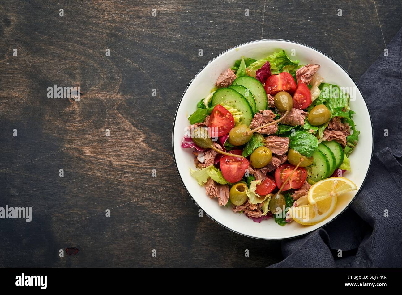Salade de thon avec légumes frais, olives, câpres et citron servie dans un bol sur fond de bois ancien. Vue de dessus avec espace de copie. Banque D'Images Salade de thon avec légumes frais, olives, câpres et citron servie dans un bol sur fond de bois ancien. Vue de dessus avec espace de copie. Banque D'Images