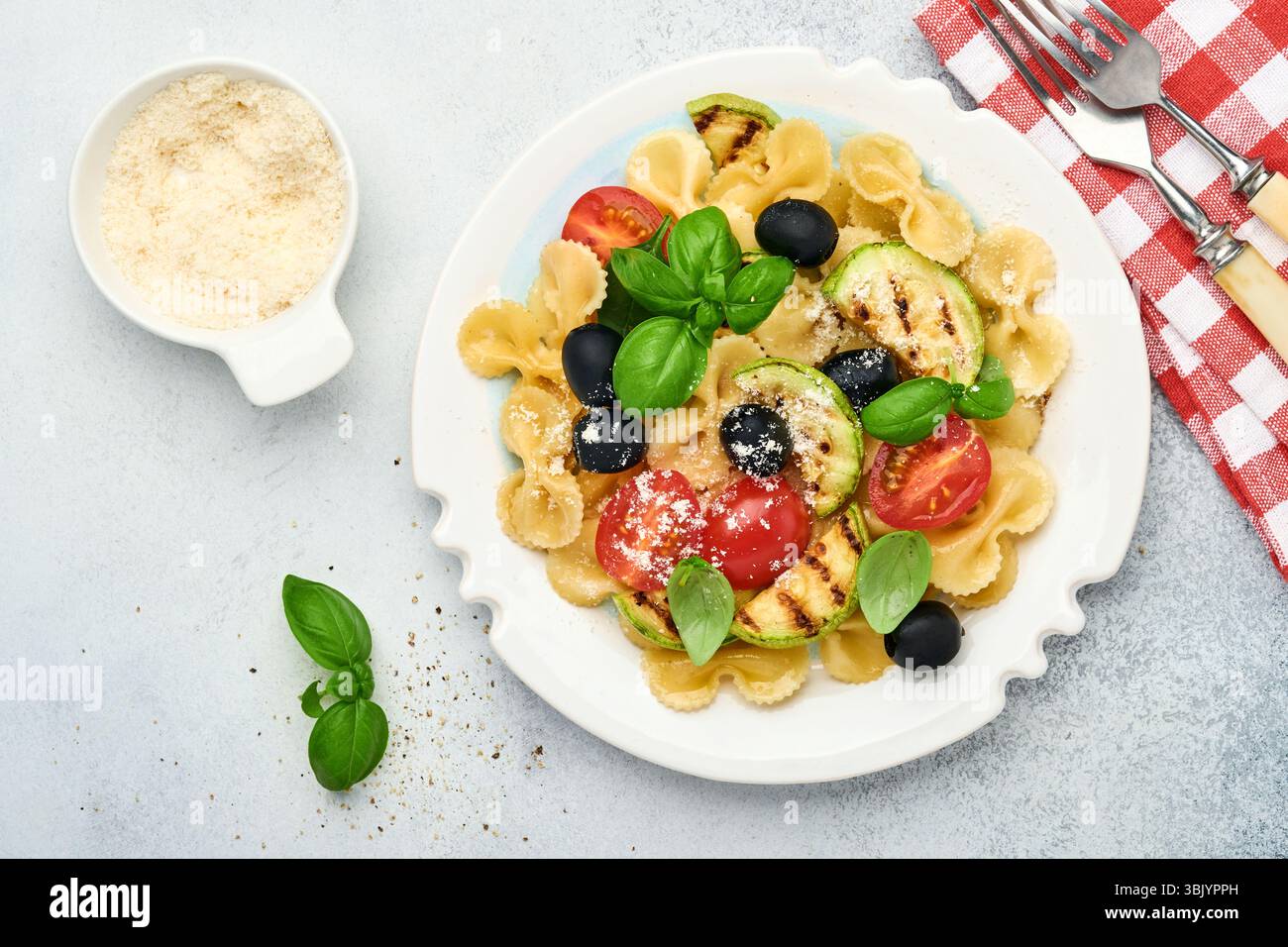 Salade de pâtes fiocchi avec légumes courgettes grillées, tomates cerises, olives, basilic et fromage parmesan dans une assiette blanche sur lumière s. Banque D'Images