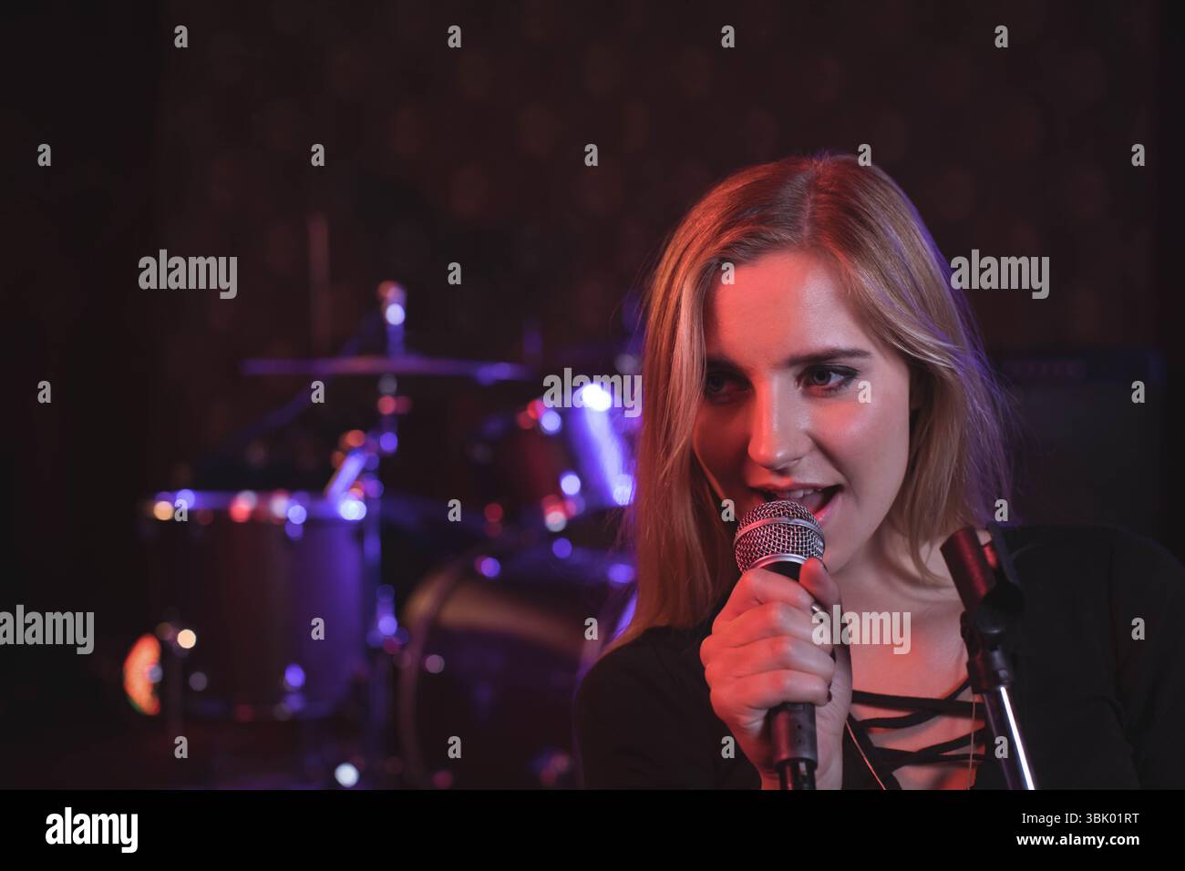 Femme tenant le microphone tout en chantant sur la petite scène à côté du kit de batterie sous les lumières rouges, copiez l'espace Banque D'Images