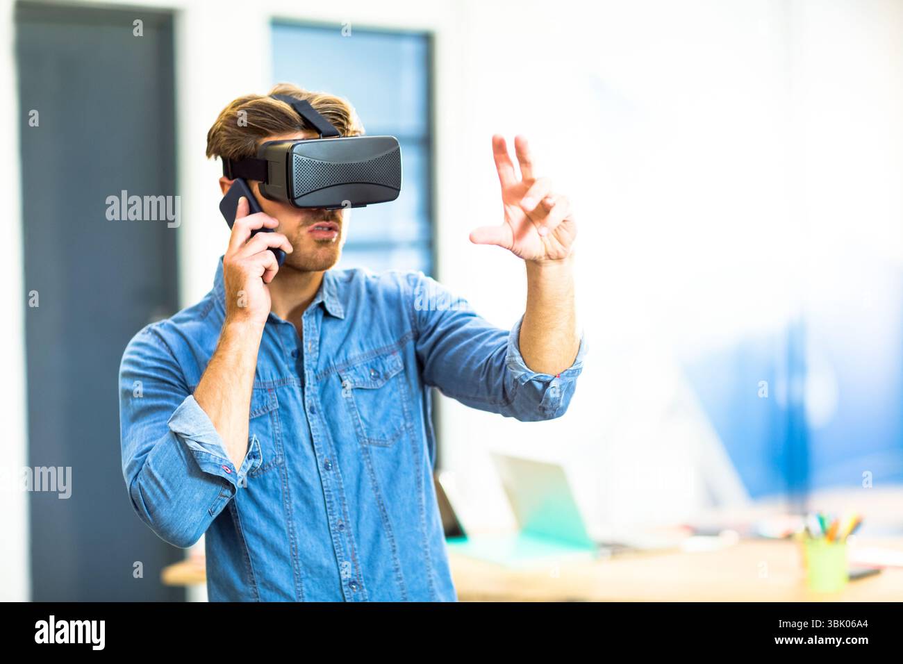 Le professionnel de la technologie porte un casque VR et tient un smartphone à côté du bureau avec un ordinateur portable dans le bureau Banque D'Images