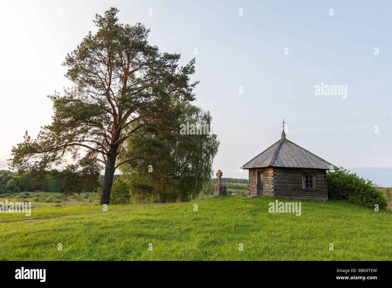 Une ancienne chapelle en bois pittoresque entourée de verdure luxuriante et de charme rustique, située dans un paysage de campagne serein avec un grand arbre dans le foreg Banque D'Images