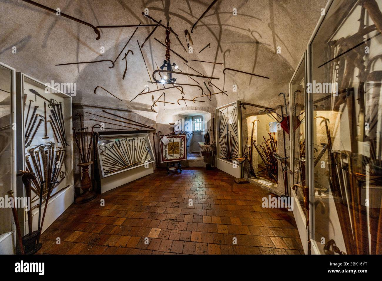 Une pièce entière remplie de cannes à marche fait partie des collections du Château de Boussac, dans le département de la Creuse, en Nouvelle-Aquitaine, France. De nombreuses cannes aux poignées sculptées et courbées sont exposées dans des vitrines et des armoires en verre. Des cannes sont accrochées en forme de éventail sur les murs et au plafond voûté et plâtré. Un luminaire est suspendu au centre, et le sol est recouvert de tuiles en briques rouges. À l'arrière, une arche ronde donne sur une pièce adjacente avec une fenêtre, devant laquelle se trouve un textile encadré sur un support. Le château est une forteresse médiévale avec une riche histoire. Le maréchal Jean de Brosse, un compagnon de Banque D'Images