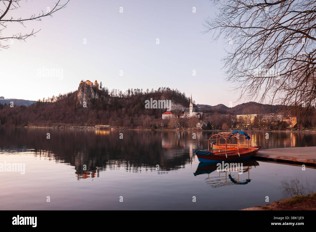 Vue au crépuscule sur le lac de Bled en Slovénie avec un château de Bled incandescent sur sa falaise et la flèche de l'église Saint-Martin reflétée dans l'eau calme, tandis qu'un pletna vide Banque D'Images