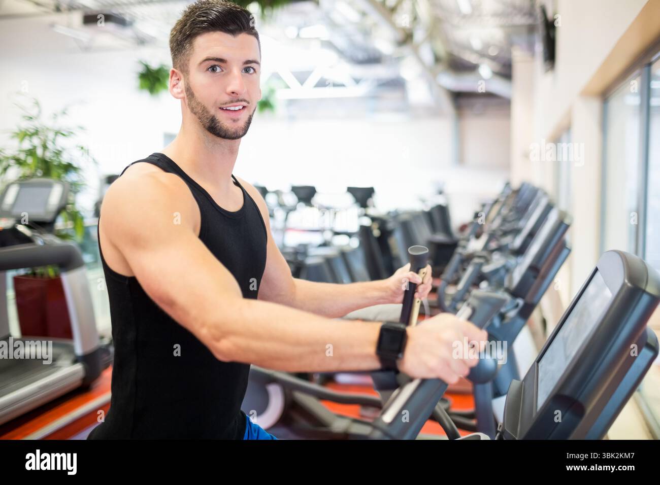 Masculin portant smartwatch s'exerçant sur un entraîneur elliptique dans la zone cardio avec des tapis roulants par les fenêtres Banque D'Images