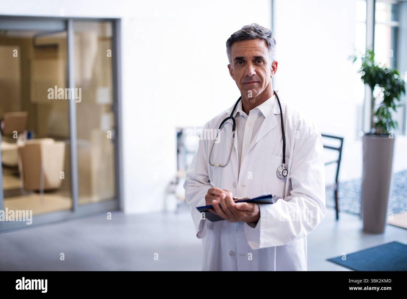 Médecin avec stéthoscope écrit des notes sur le presse-papiers près de la cloison en verre et des chaises beiges Banque D'Images