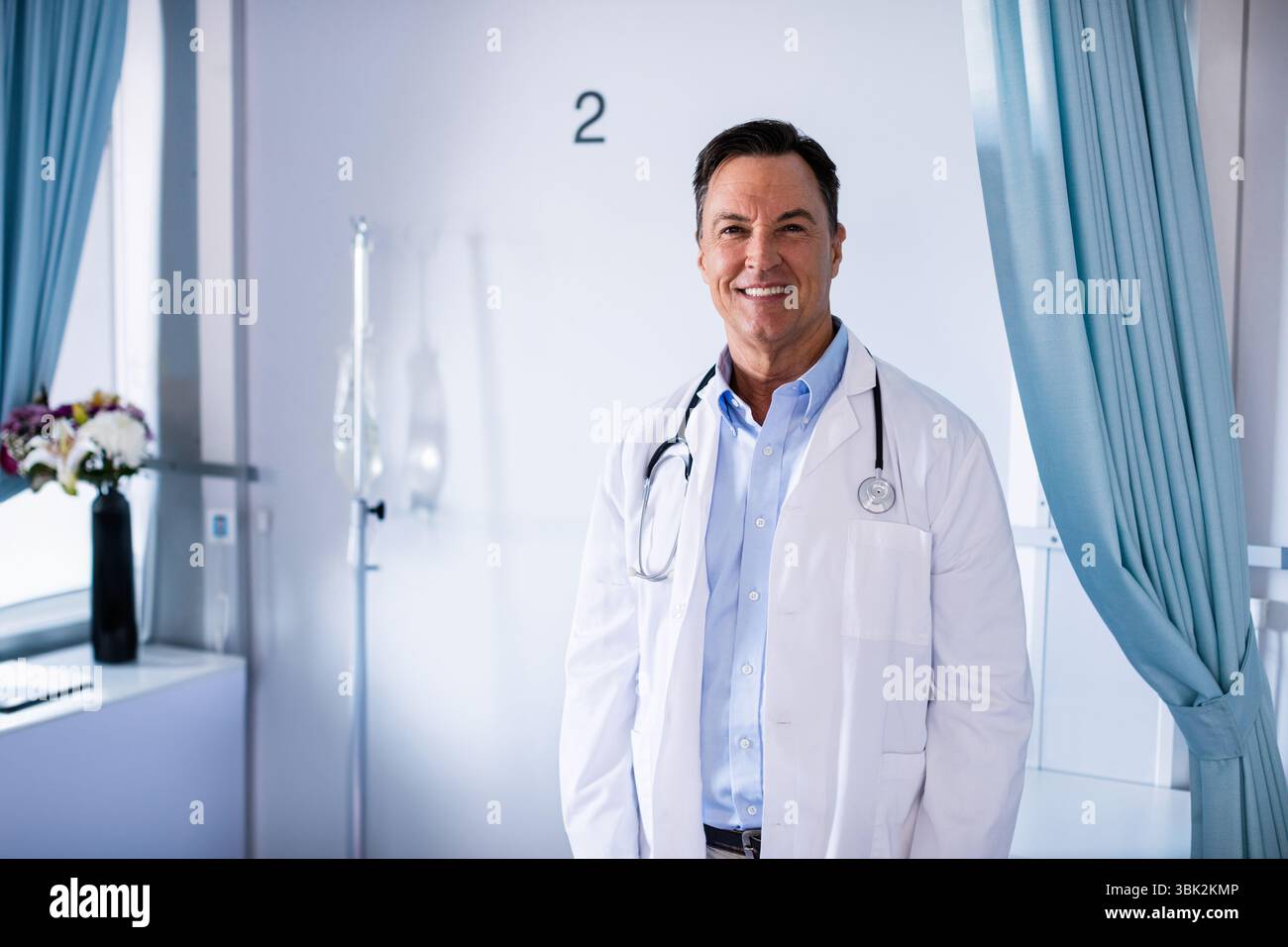 Médecin portant un stéthoscope debout dans la baie de l'hôpital 2 à côté de la perche IV et des fleurs près des rideaux bleus Banque D'Images
