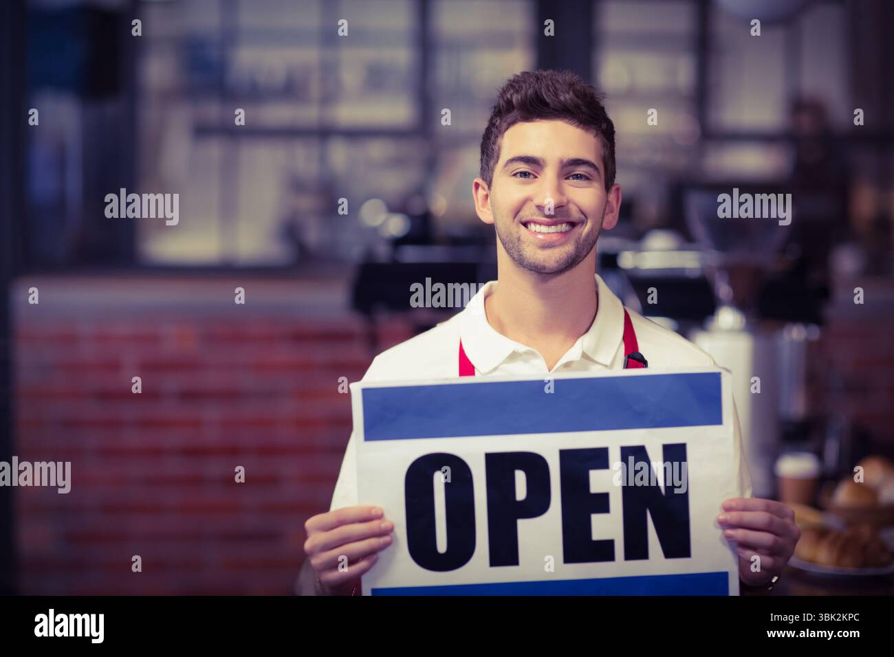 Barista portant une chemise blanche et un tablier rouge portant un panneau OUVERT bleu et blanc au comptoir du café en briques Banque D'Images