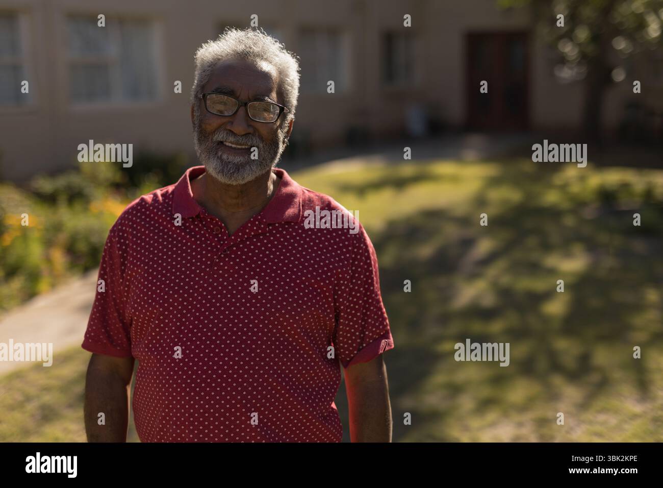 Homme senior afro-américain debout dans la cour avant ensoleillée portant un polo rouge et des lunettes Banque D'Images