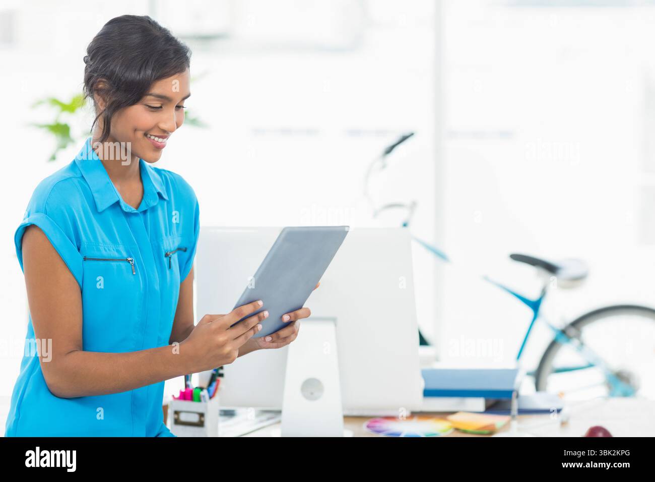 Femme sud-asiatique debout au bureau tenant une tablette parmi la papeterie, le vélo et la plante près des fenêtres Banque D'Images